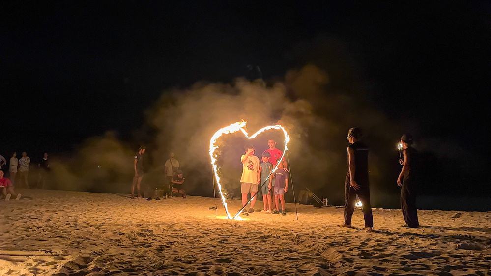 A group of people are standing on a beach at night, surrounded by a heart-shaped fire formation.