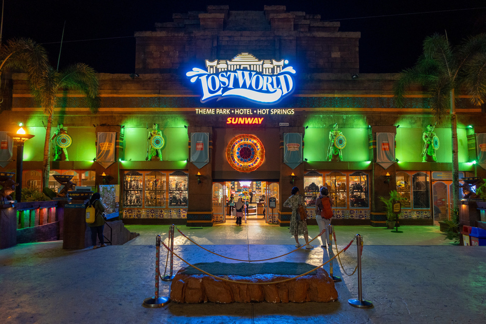 The photo shows the illuminated entrance of the Lost World Theme Park with people entering and vibrant green and red lights.