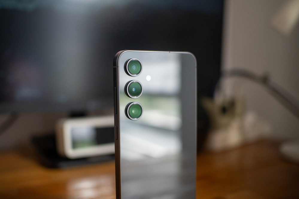 A smartphone with three rear cameras is positioned on a table, with a blurred background featuring a monitor and other objects.