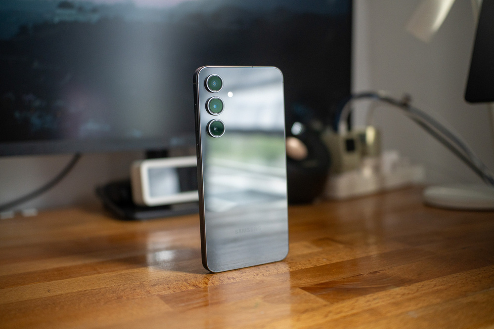 A smartphone with a reflective back and three camera lenses is standing upright on a wooden desk.
