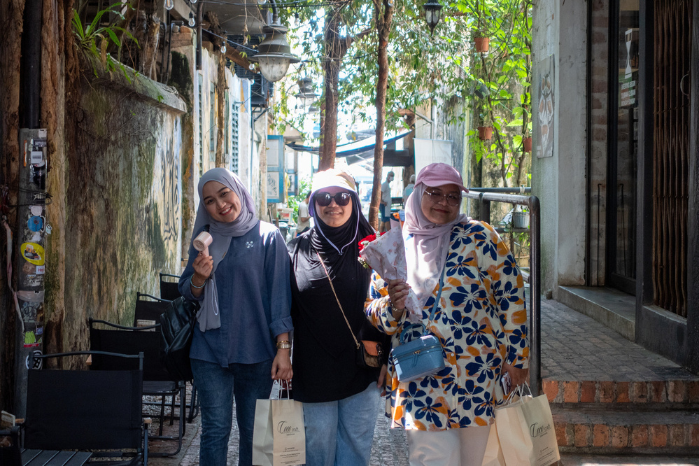 Three people are walking along a narrow alley holding shopping bags and drinks, with trees and buildings on either side.