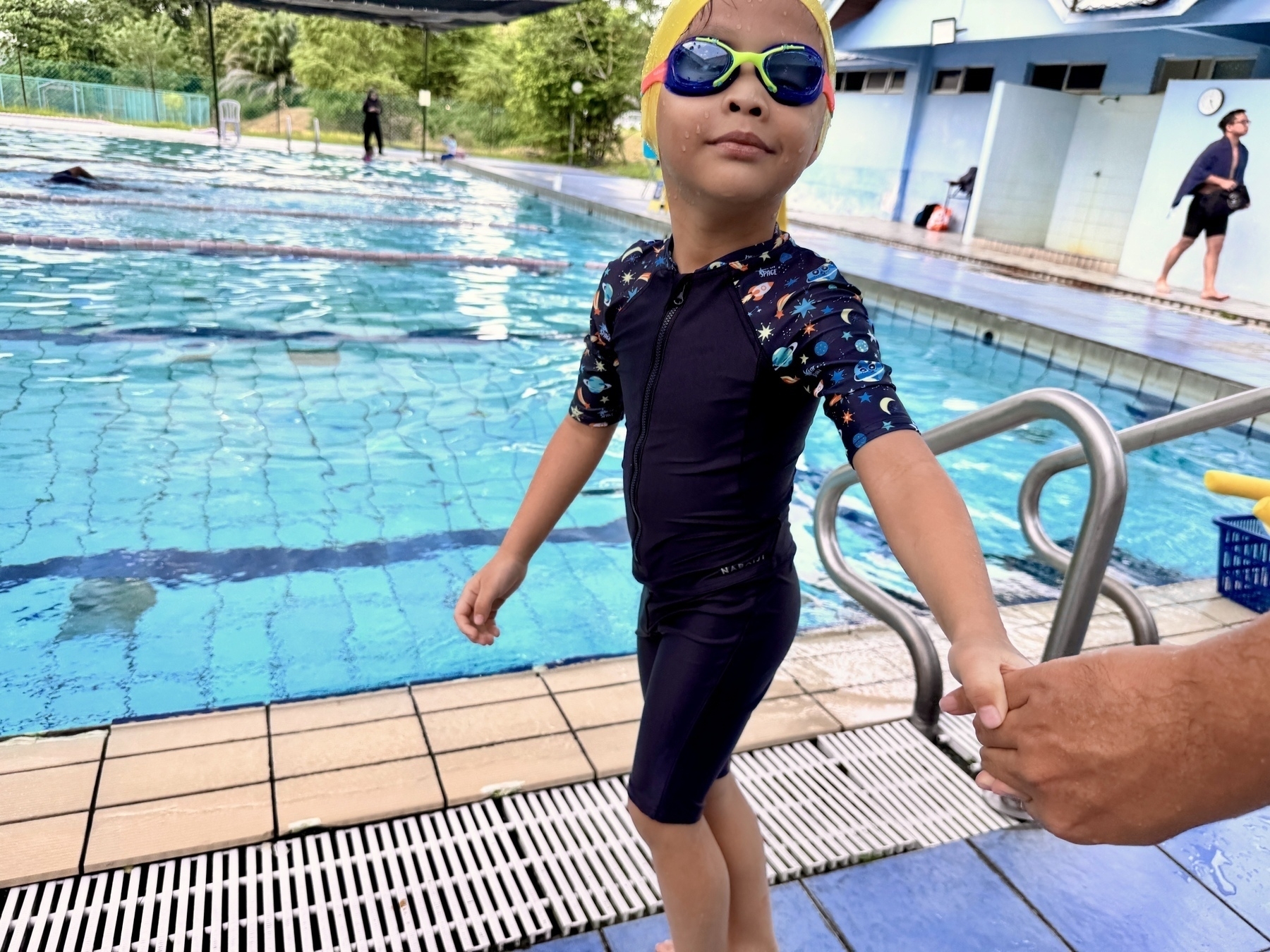 A child wearing swim gear and goggles holds an adult's hand beside a swimming pool.