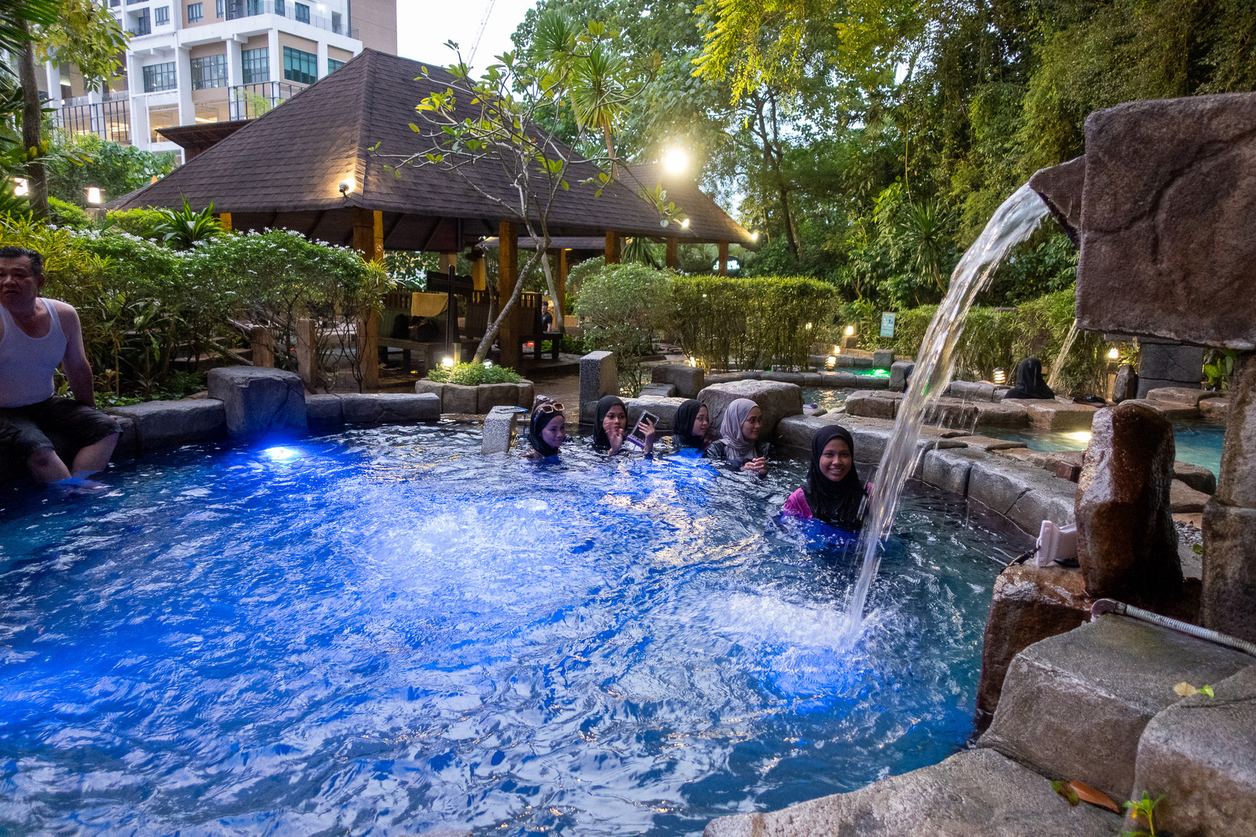 A group of people enjoy a swim in a pool with a waterfall feature, surrounded by lush greenery and illuminated by soft blue lighting.
