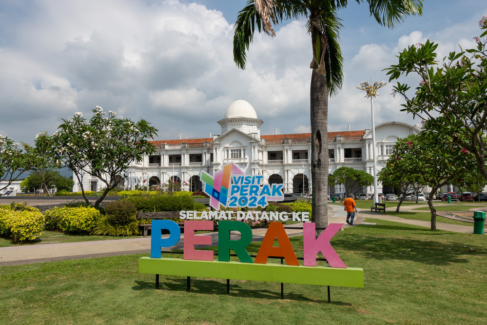 A large sign welcoming visitors to Perak stands in a park with a grand building in the background under a partly cloudy sky.