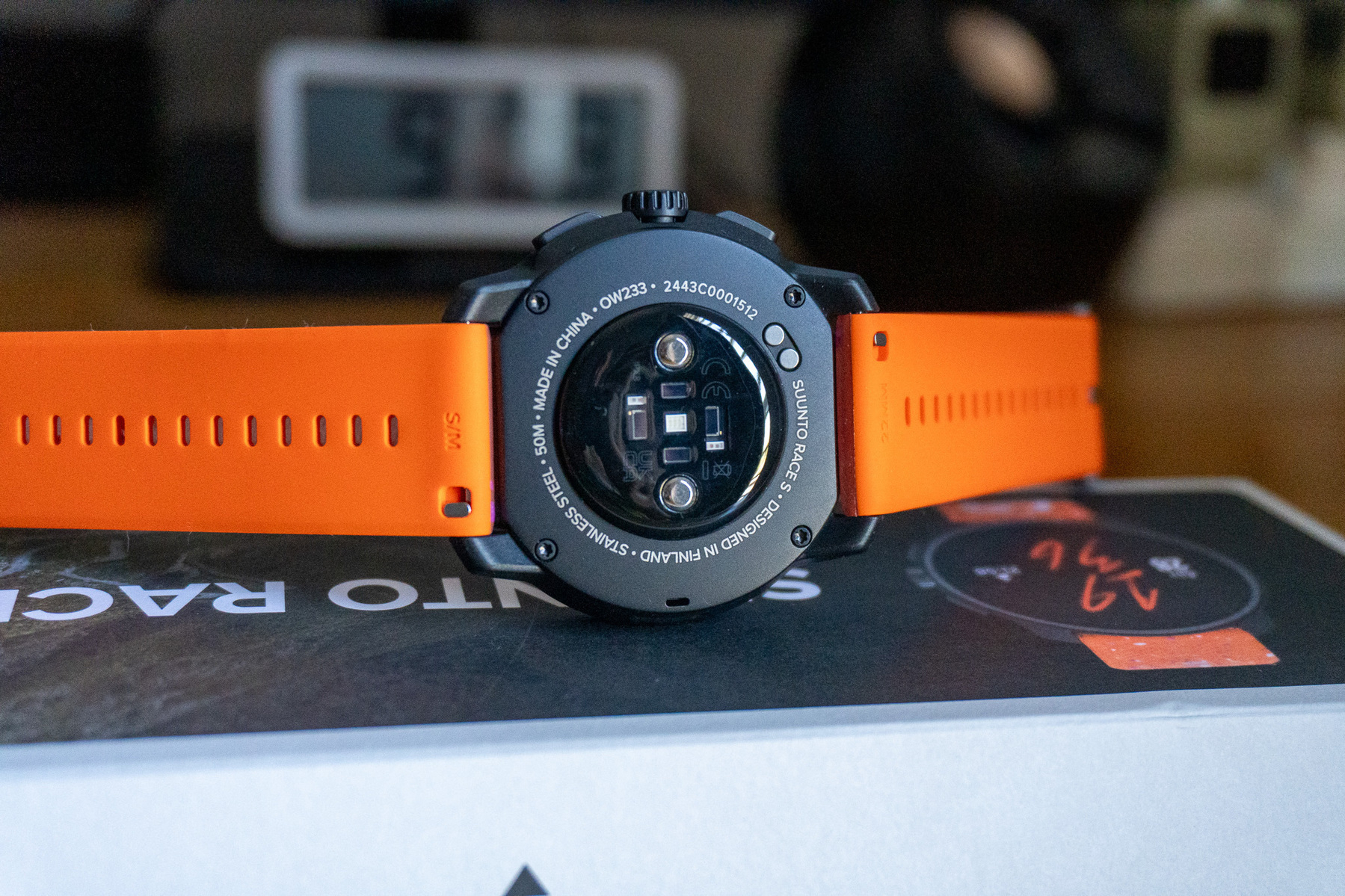 A smartwatch with an orange band is placed face down on a box, revealing its sensors and back cover details.