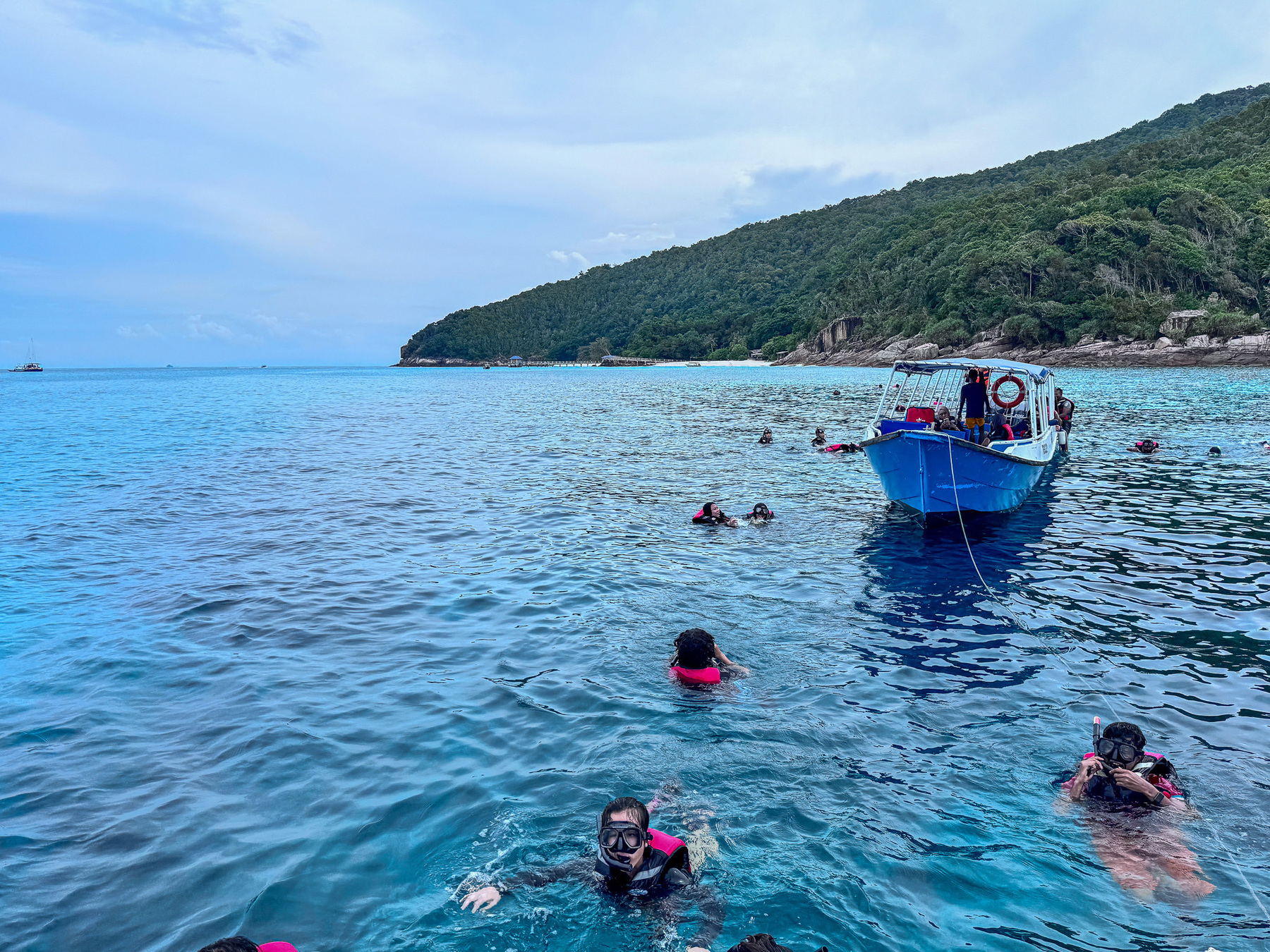 Auto-generated description: A group of snorkelers wearing life vests is swimming near a small boat in clear blue waters with a forested island in the background.
