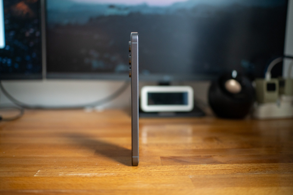 A smartphone is standing on its edge on a wooden desk, surrounded by electronic devices and cables in the background.