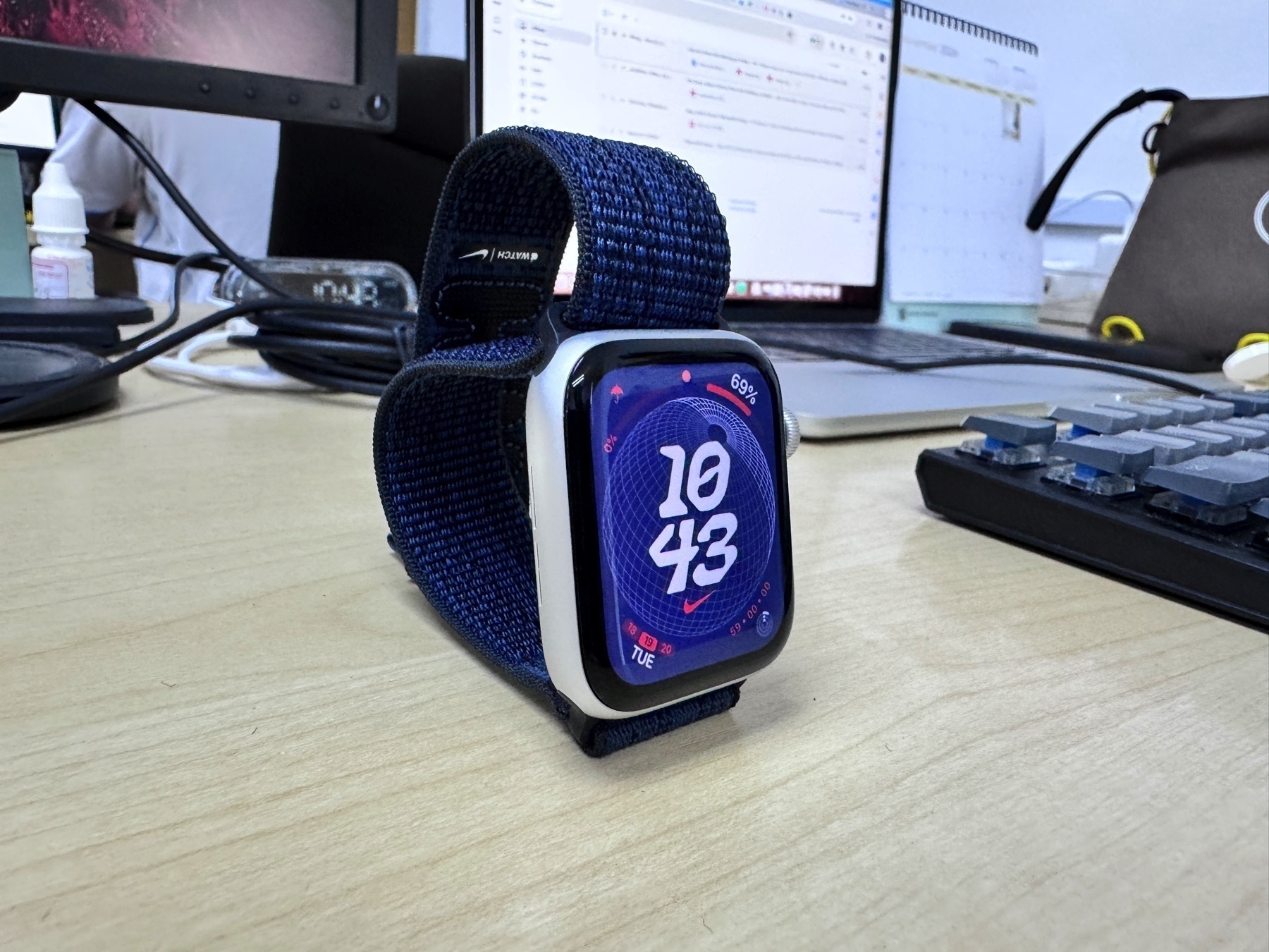 A smartwatch with a blue strap showing the time is displayed on a desk near a laptop and keyboard.