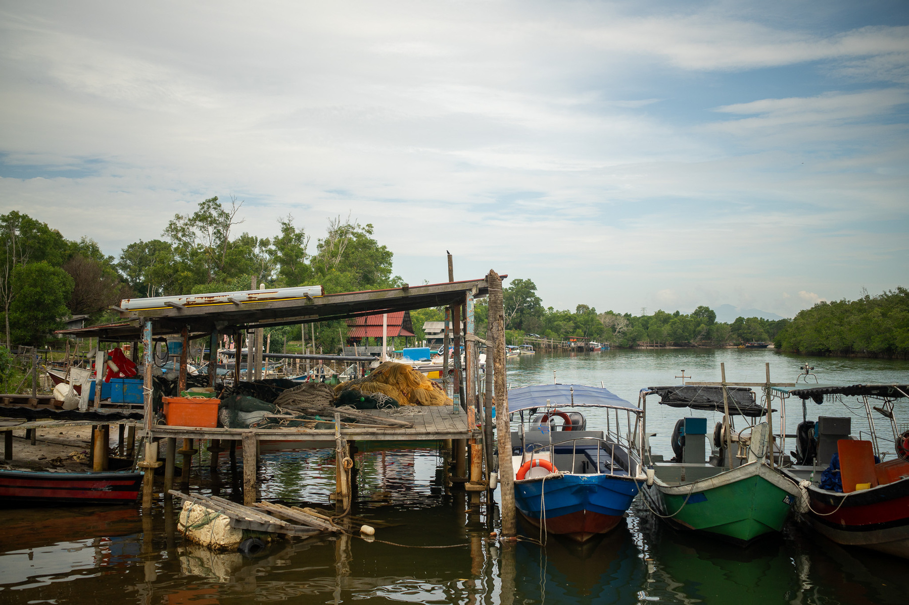 Auto-generated description: A small dock with several boats and a rustic wooden structure sits by a calm, tree-lined river.