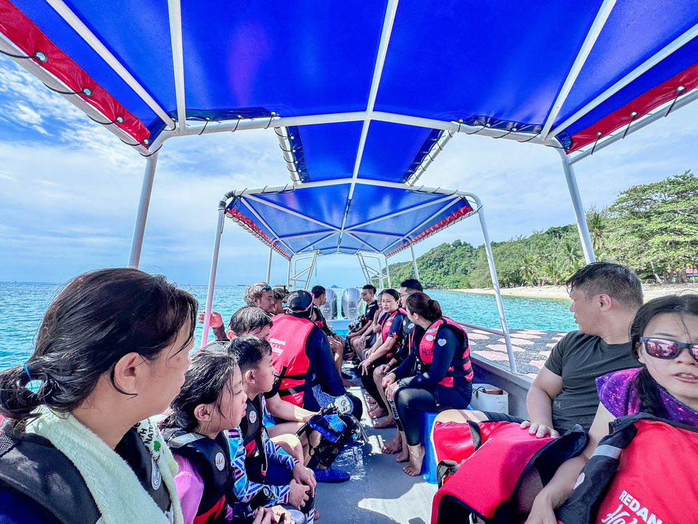 A group of people wearing life jackets are sitting on a boat with a canopy, traveling on a bright, scenic body of water near a forested coastline.
