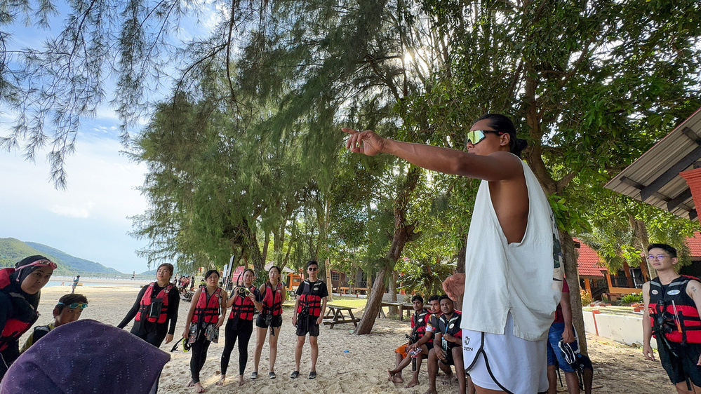 A group of people wearing life jackets listens to a person giving instructions on a beach under trees.