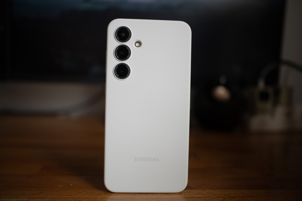 A white Samsung smartphone with three rear cameras is placed upright on a wooden surface.