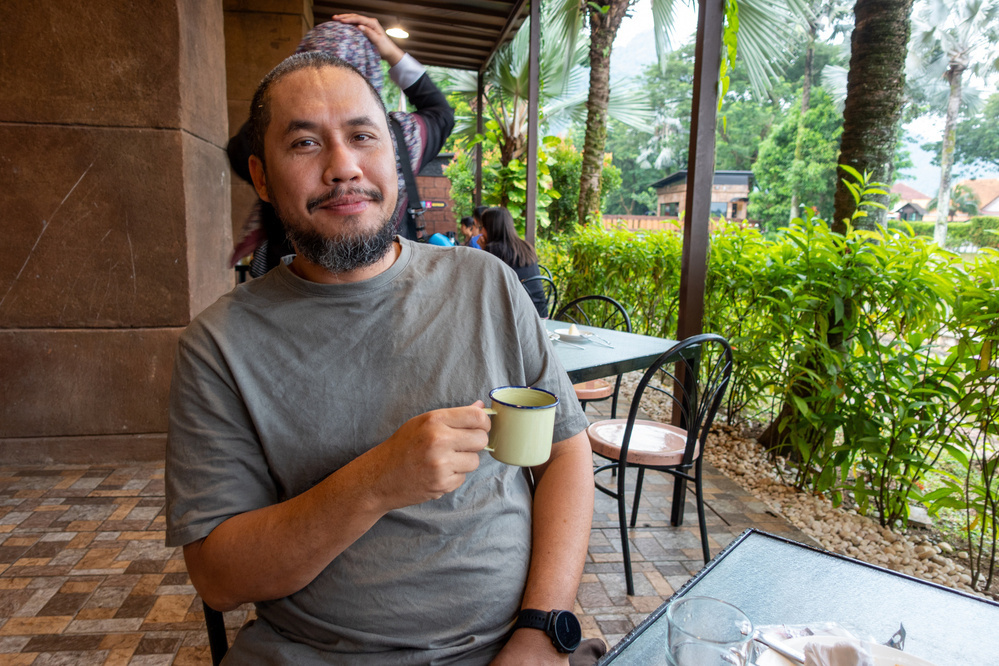 A man sits at an outdoor café table, holding a mug and smiling, with greenery in the background.