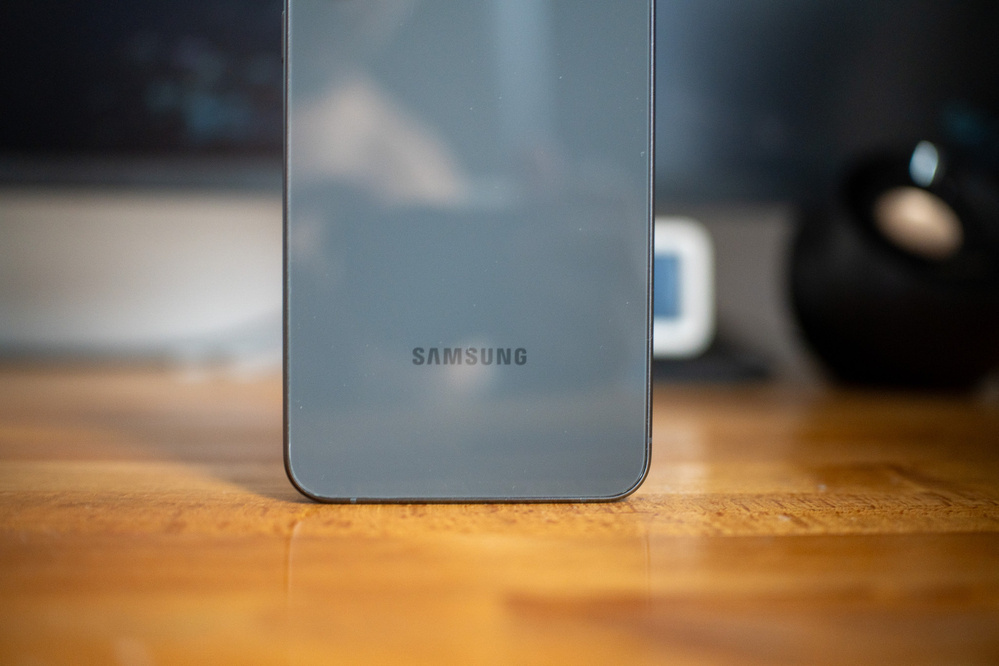 A Samsung smartphone is standing on a wooden surface with a blurred background.