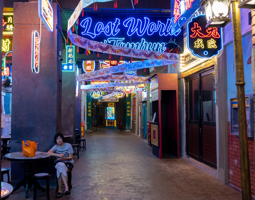 A person sits at a table in a neon-lit alley with colorful signs and decorations.