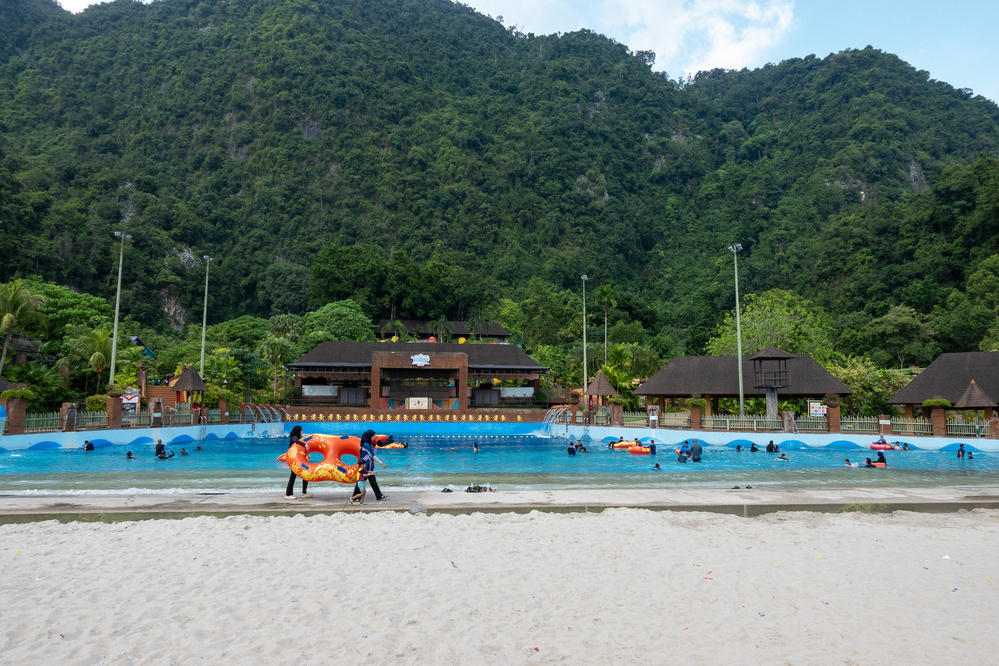 A beach scene features people enjoying a wave pool with lush green hills in the background.