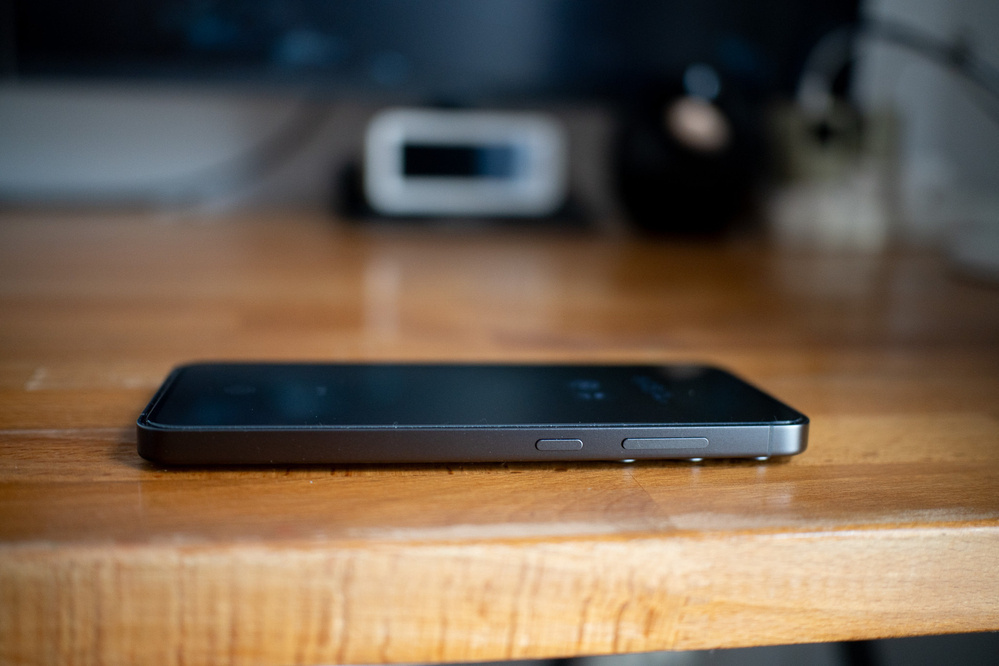 A smartphone lies face-up on a wooden table, focusing on its side buttons.