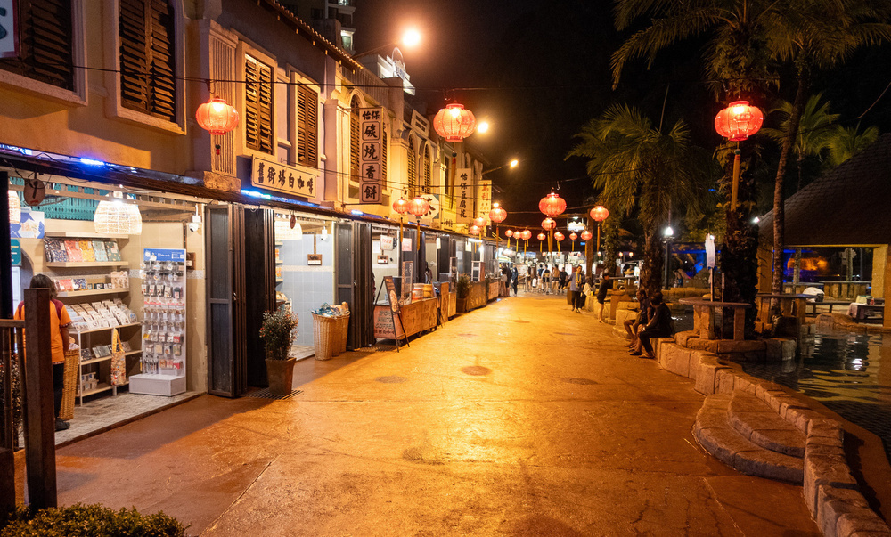 A warmly lit street at night features traditional buildings adorned with glowing red lanterns and people strolling along the sidewalk.