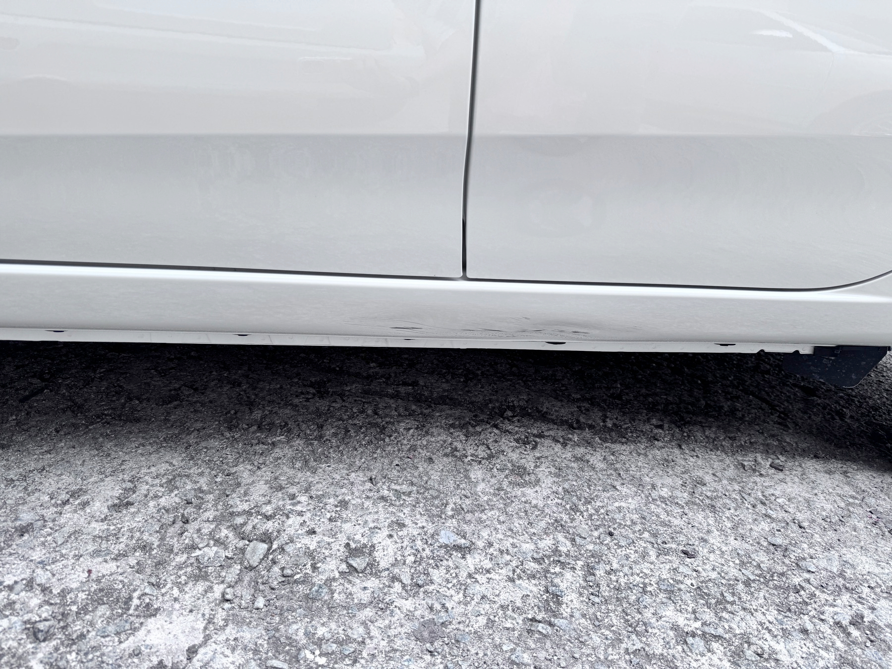 A white car parked on a rough, gravelly surface with the focus on its lower side panel.