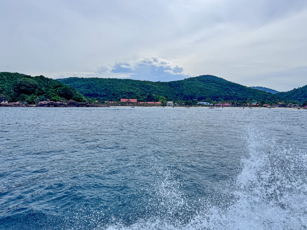 A waterfront landscape features a vast body of water with waves in the foreground and a lush, mountainous shoreline in the background under a cloudy sky.