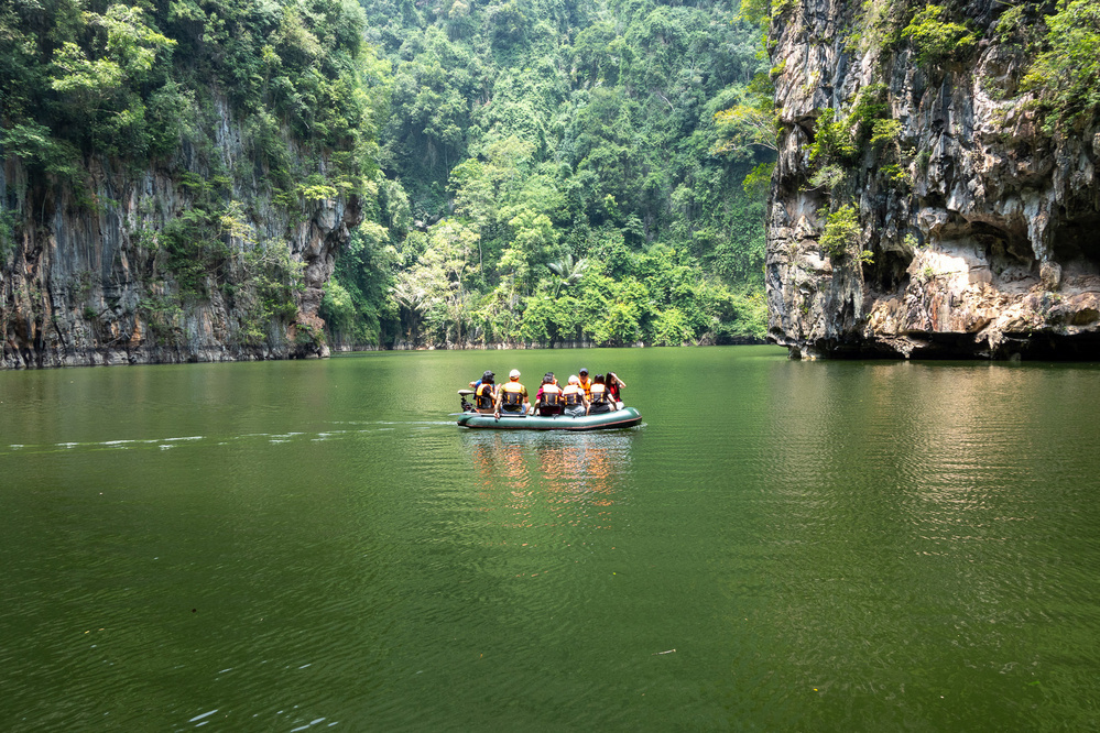 A group of people wearing life jackets are in a small boat on a green lake surrounded by lush, rocky cliffs.