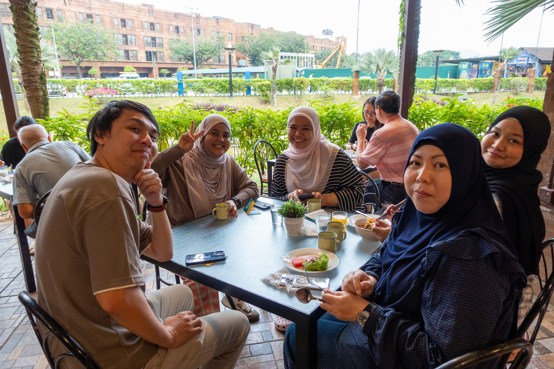 A group of people is seated around a table outdoors, enjoying food and drinks, with greenery and buildings in the background.