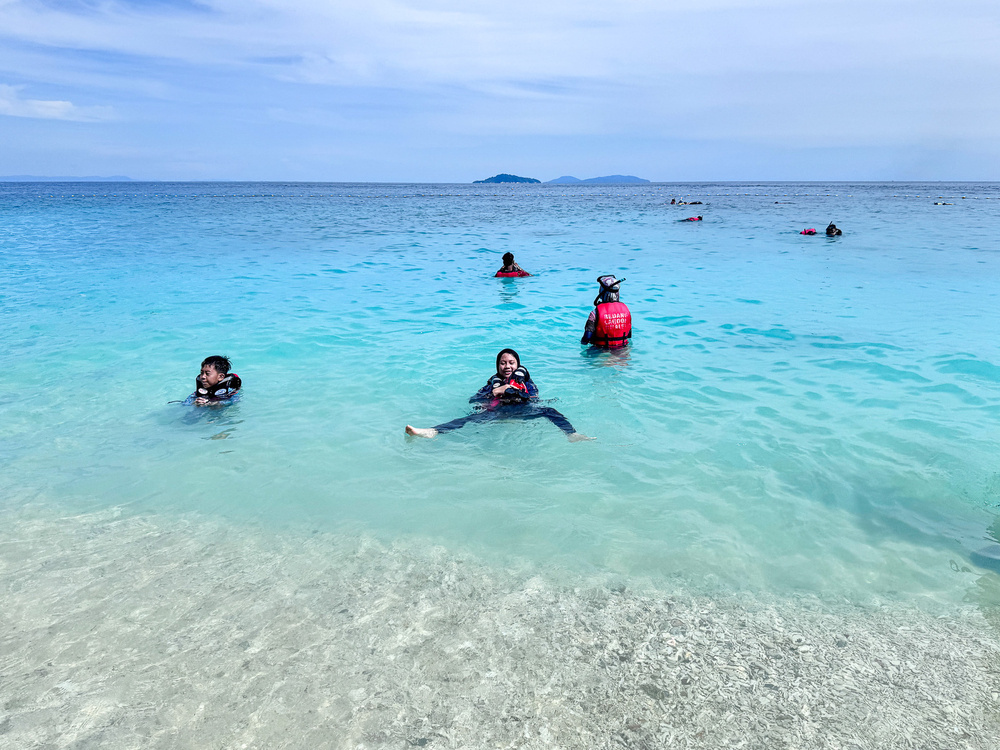 People are enjoying swimming and floating in clear blue ocean water near a sandy beach.