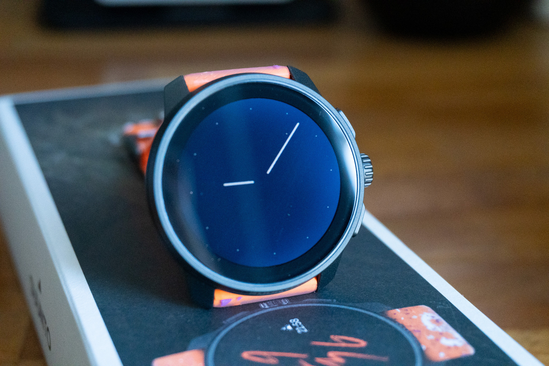 A smartwatch with a minimalist analog-style face and an orange strap is resting on a box.
