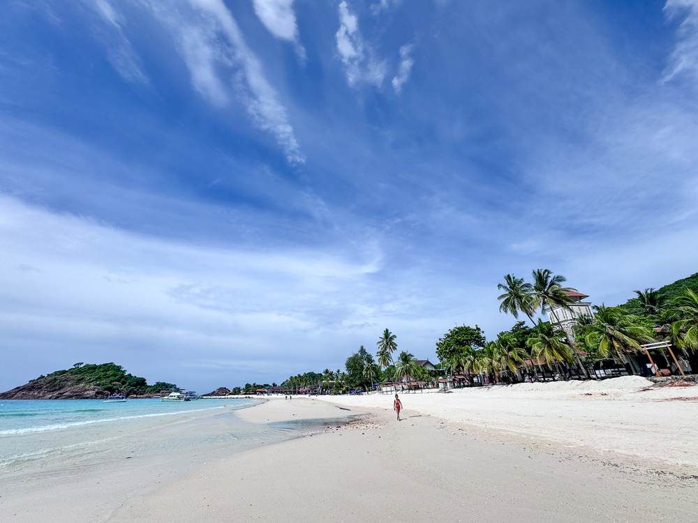 A serene beach scene features white sand, palm trees, a person walking along the shoreline, and a clear blue sky scattered with clouds.