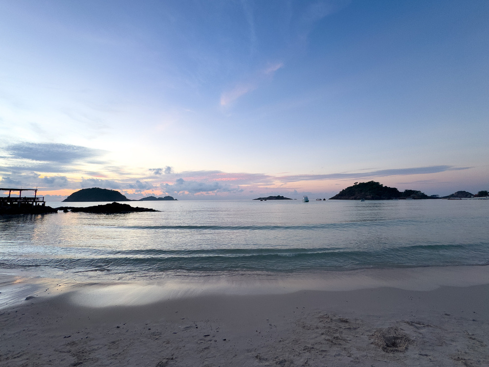 A tranquil beach scene at sunset features calm waves, a sandy shore, and distant islands under a pastel sky.