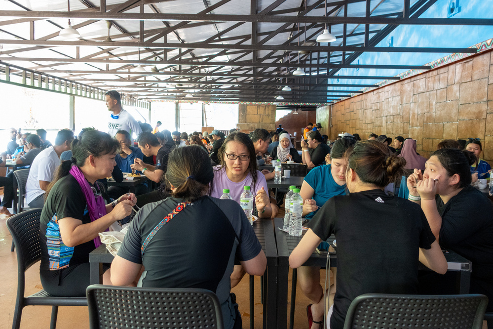 A large group of people is sitting and eating at tables in a busy, covered outdoor dining area.