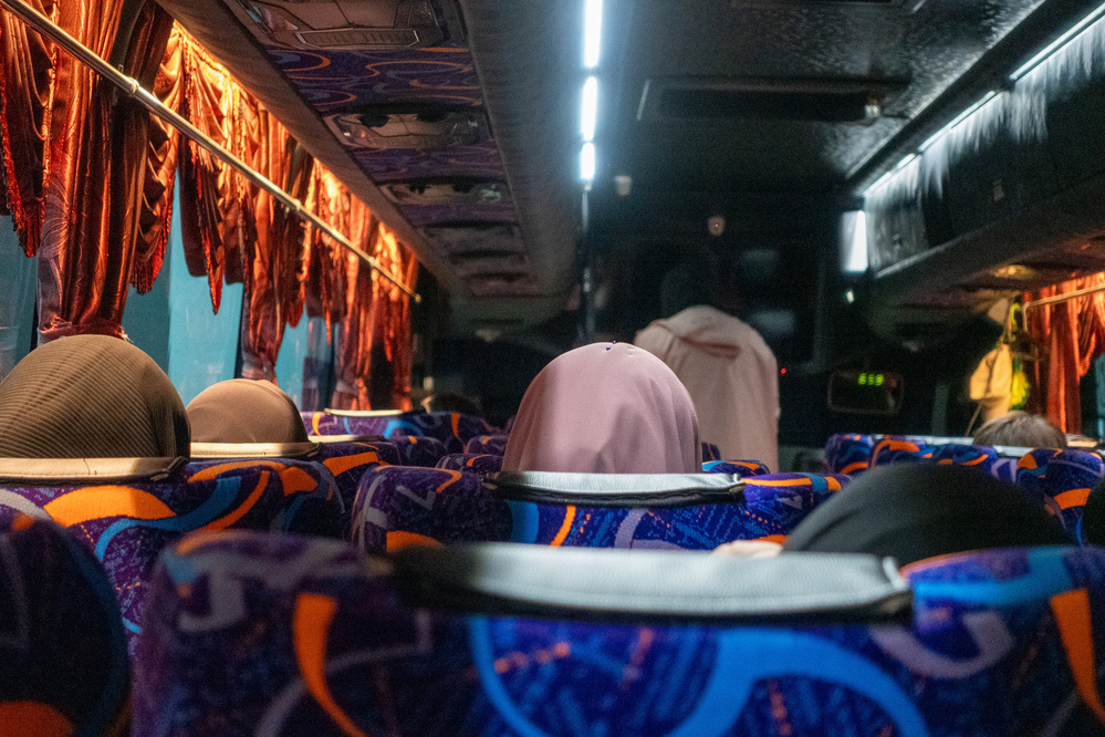 Passengers are seated inside a bus with vibrant patterned seats and red curtains.