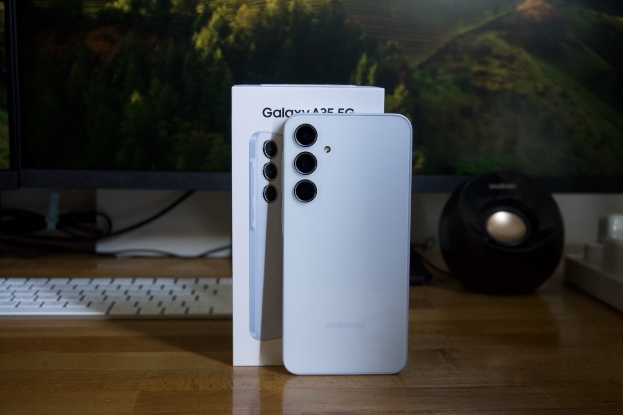 Auto-generated description: A white smartphone with three camera lenses is displayed alongside its box on a wooden surface, with a monitor and keyboard in the background.