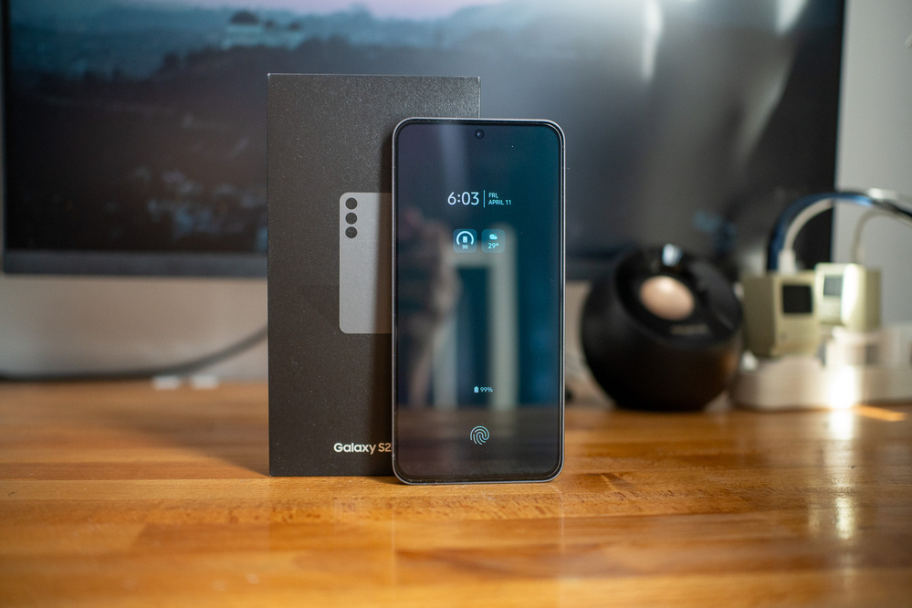 A smartphone displaying the time is propped against its box on a wooden surface, with a blurred background including a monitor and various desk items.