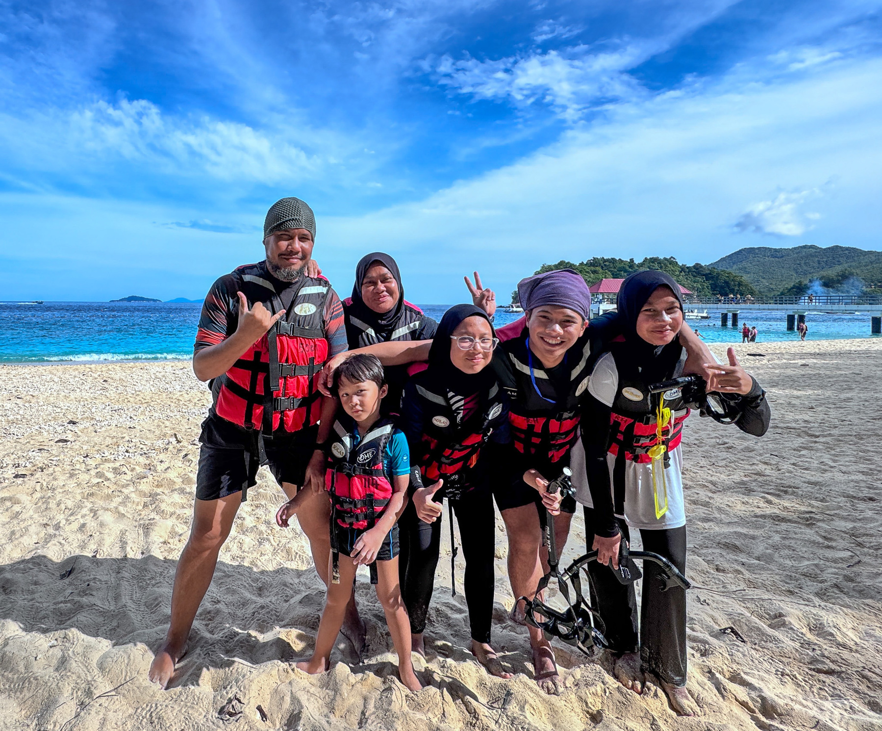 Auto-generated description: A group of six people wearing life jackets is posing happily on a sandy beach with the ocean and hills in the background.