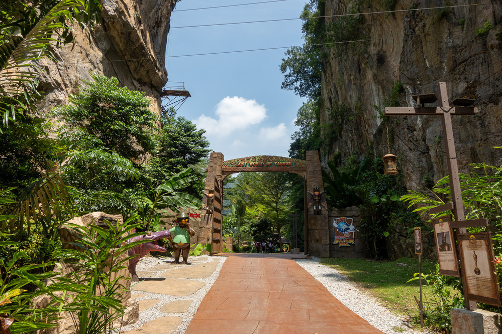 A scenic pathway leads through a stone archway flanked by towering cliffs and lush greenery under a clear blue sky.