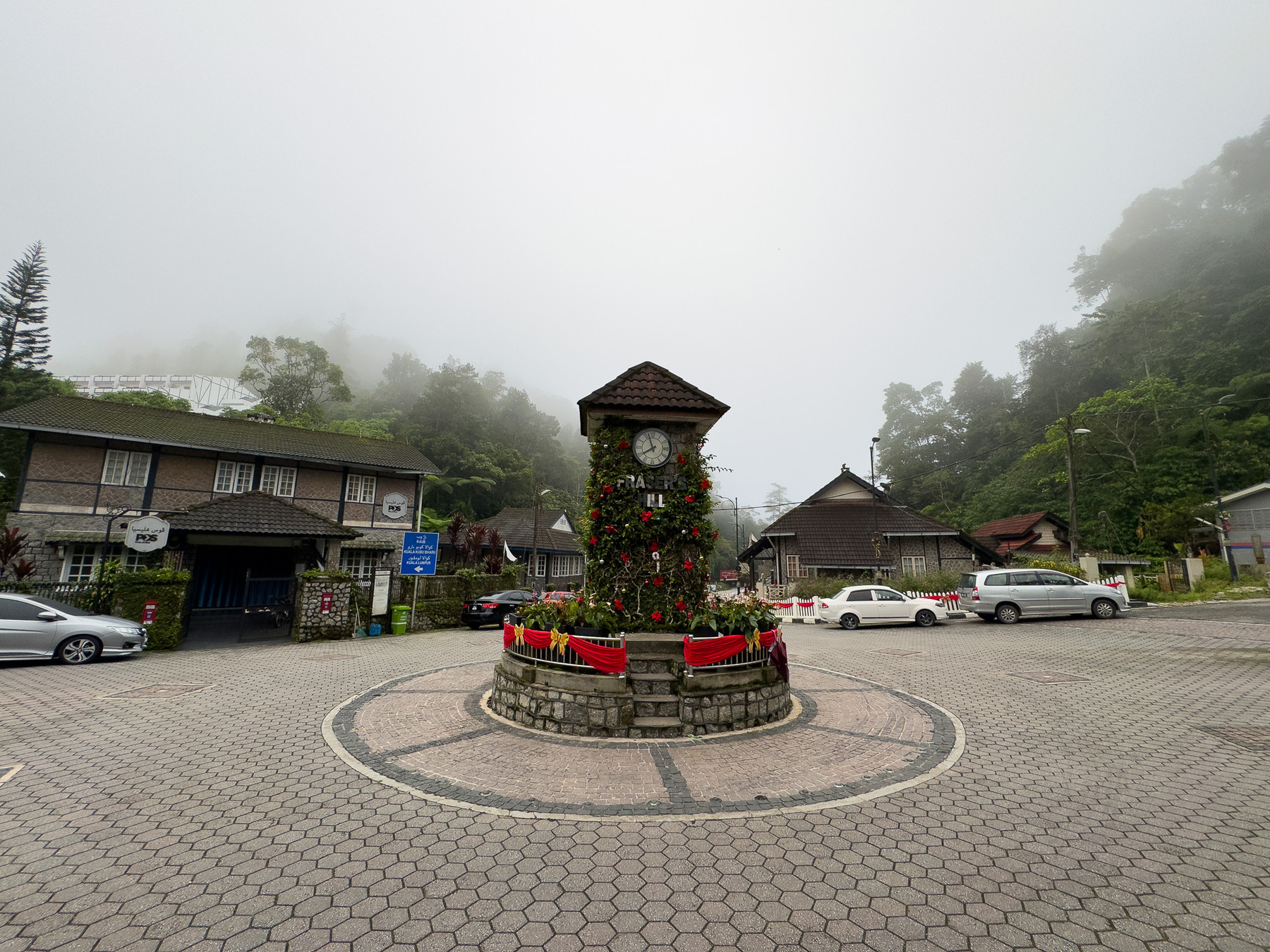 A quaint village scene features a central clock tower adorned with flowers, surrounded by mist and greenery, with buildings and parked cars nearby.