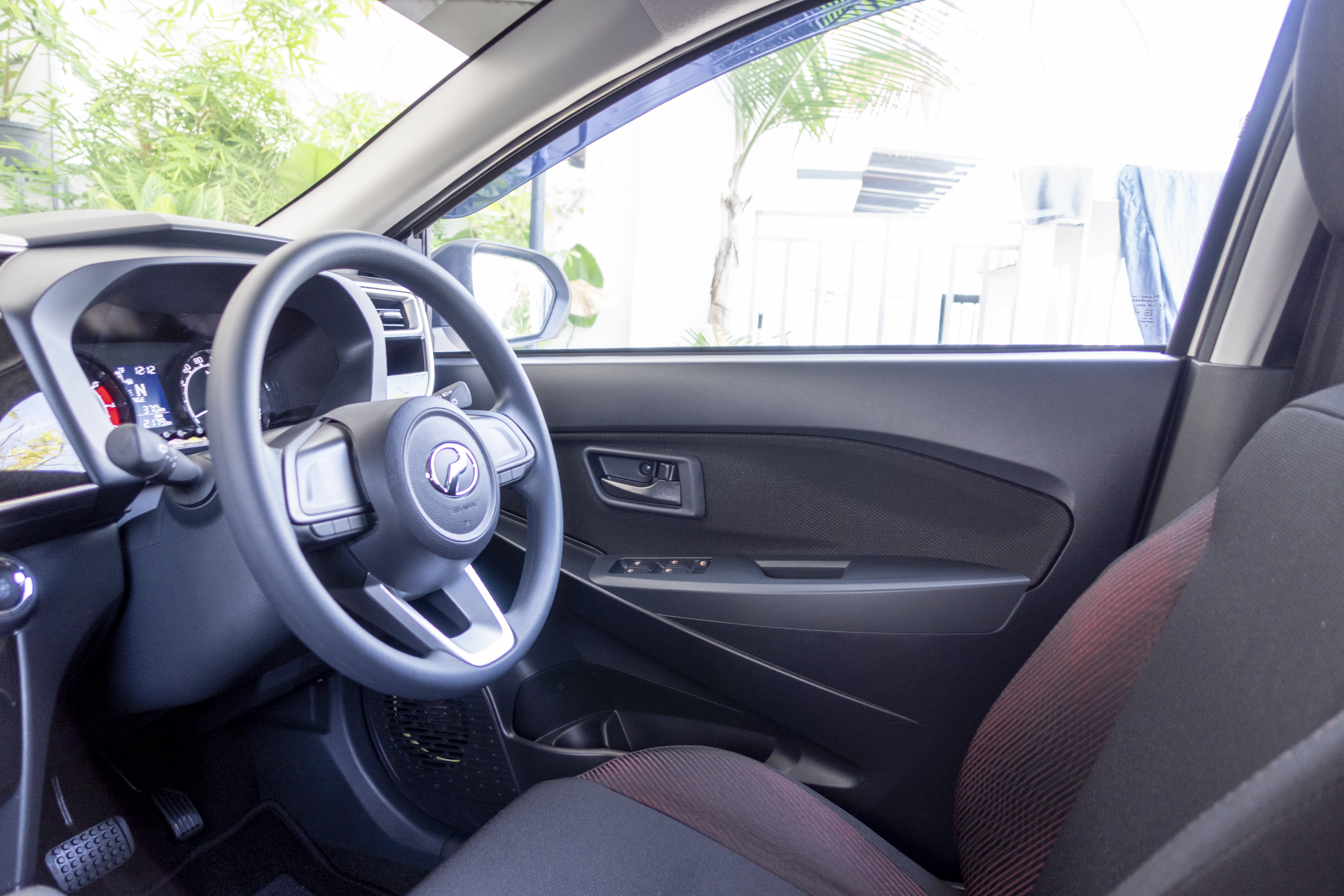 A modern car interior features a sleek dashboard, steering wheel, and driver's seat with green foliage visible through the window.