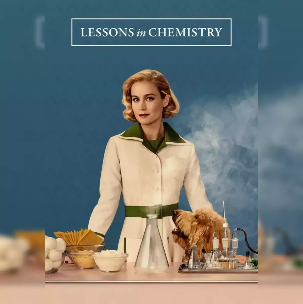 Auto-generated description: A woman stands in front of a table with chemistry lab equipment, food ingredients, and a dog, beneath the title Lessons in Chemistry.