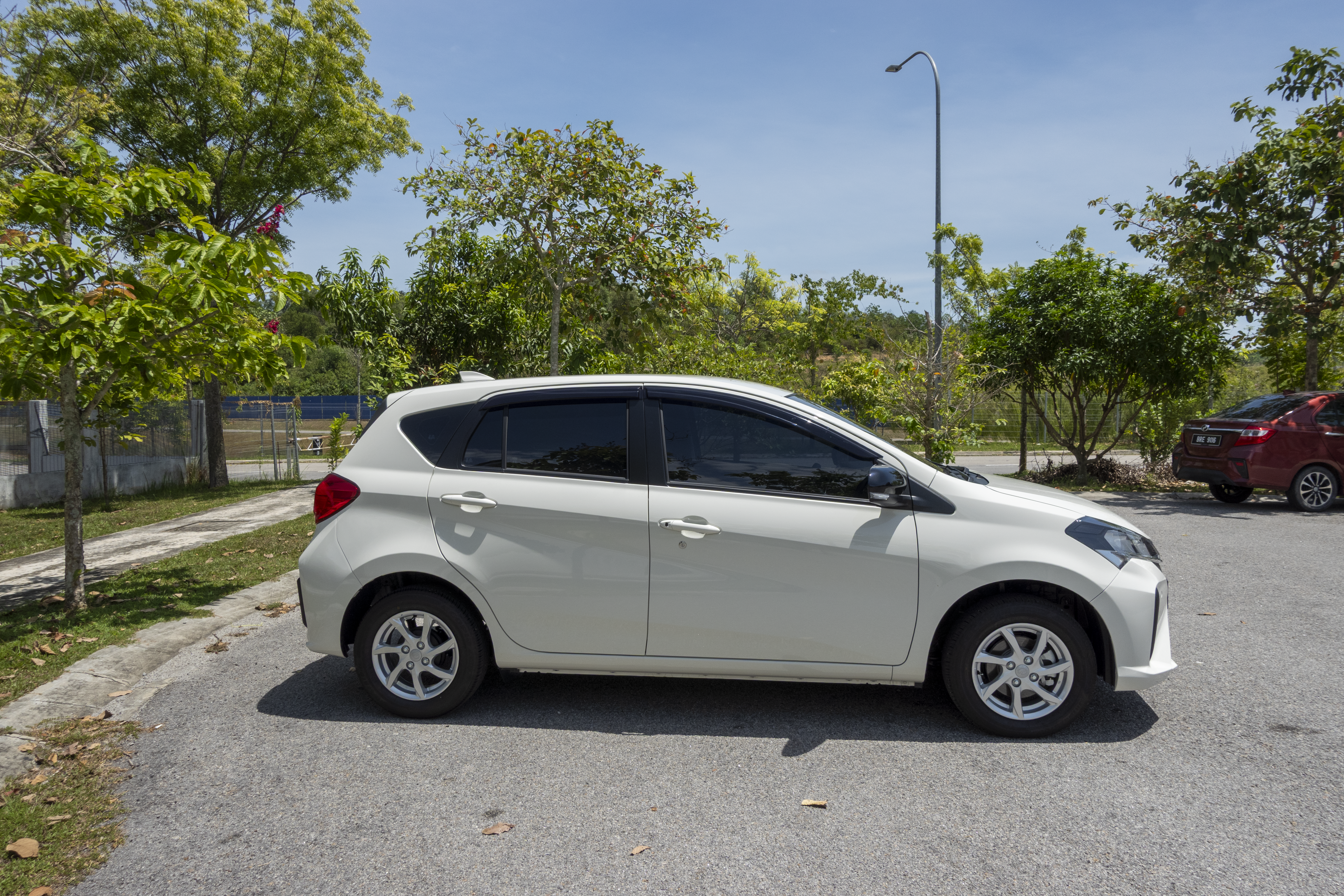 A white hatchback car is parked in a shaded area surrounded by trees.
