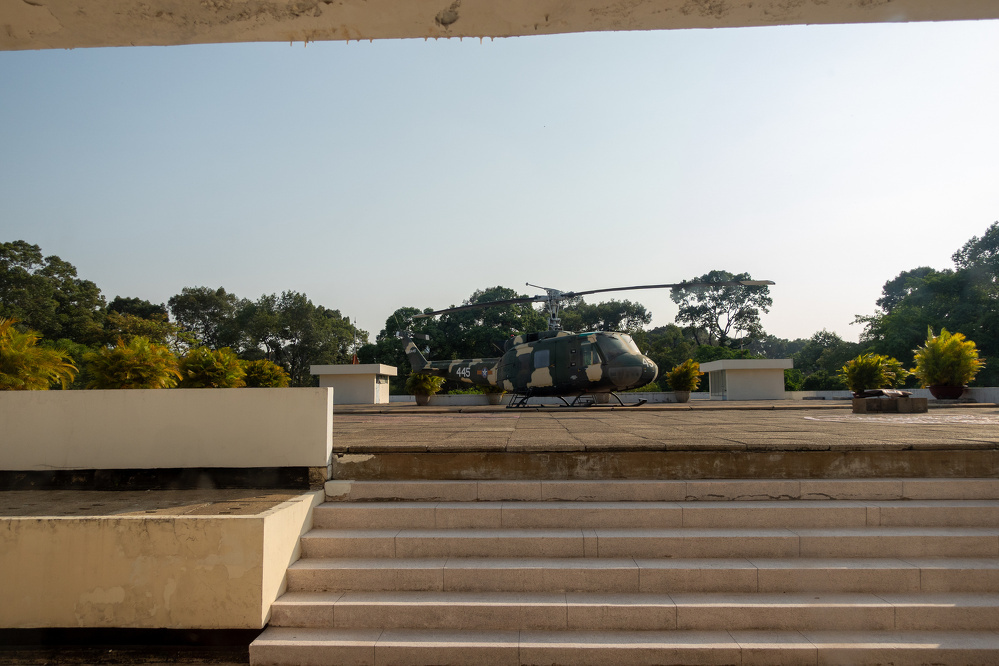 A helicopter is displayed on a platform surrounded by greenery and two white structures in the background under a clear sky.