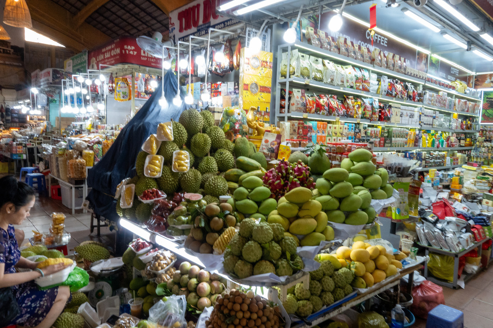 A vibrant market stall is filled with a variety of tropical fruits and packaged goods, with a person sitting and working nearby.