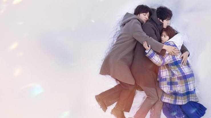 Two individuals wearing coats lie in the snow, embracing a third person wrapped in a checkered blanket, creating a scene of warmth and comfort.
