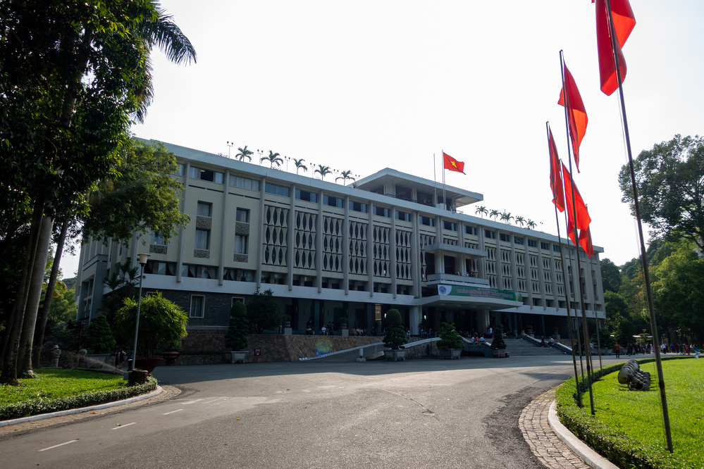 A large government or institutional building is surrounded by trees and several red flags with yellow stars are displayed prominently.