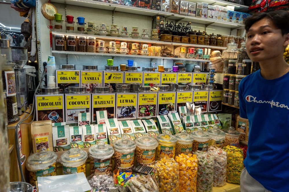 A market stall displaying various types of coffee and snacks with clear labels and a young man wearing a blue Champion shirt standing nearby.
