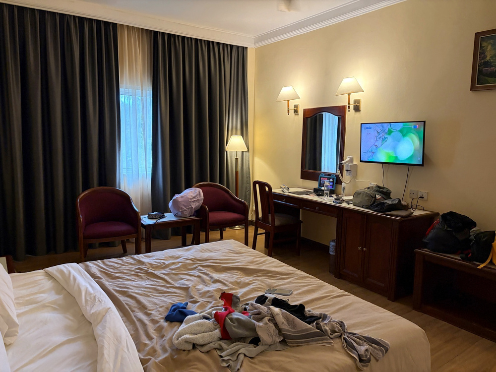 A cozy hotel room features a neatly arranged bed, a small table with chairs, a television, and a desk cluttered with personal items.