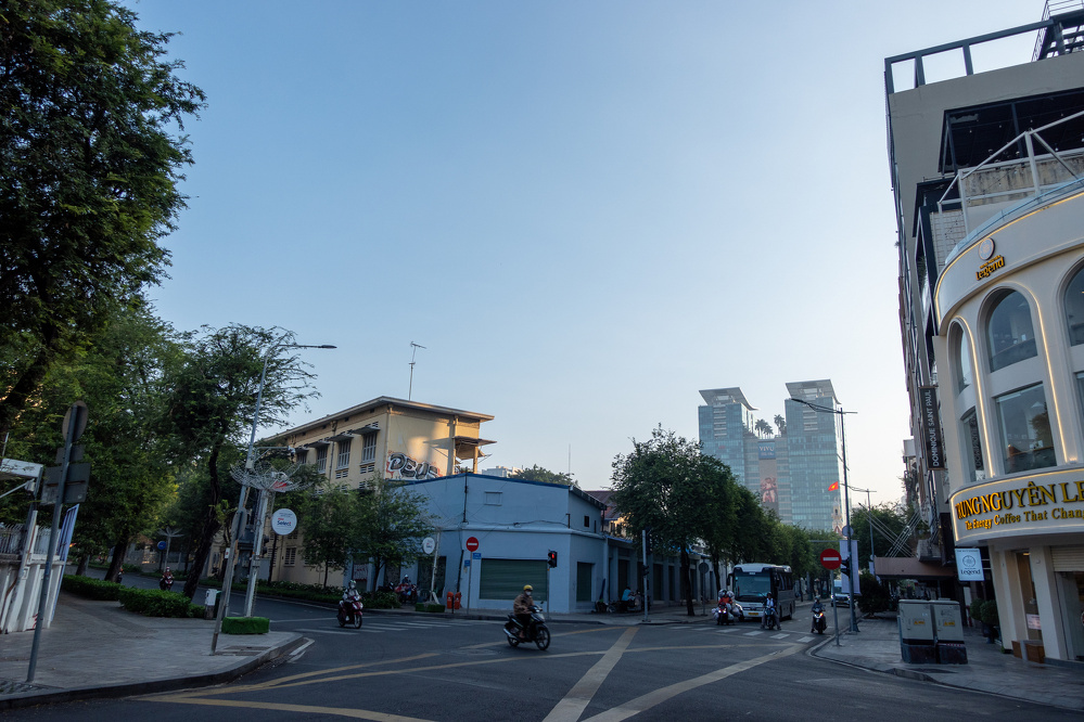 A quiet city street is lined with buildings and trees under a clear sky with a few motorcyclists riding along the road.