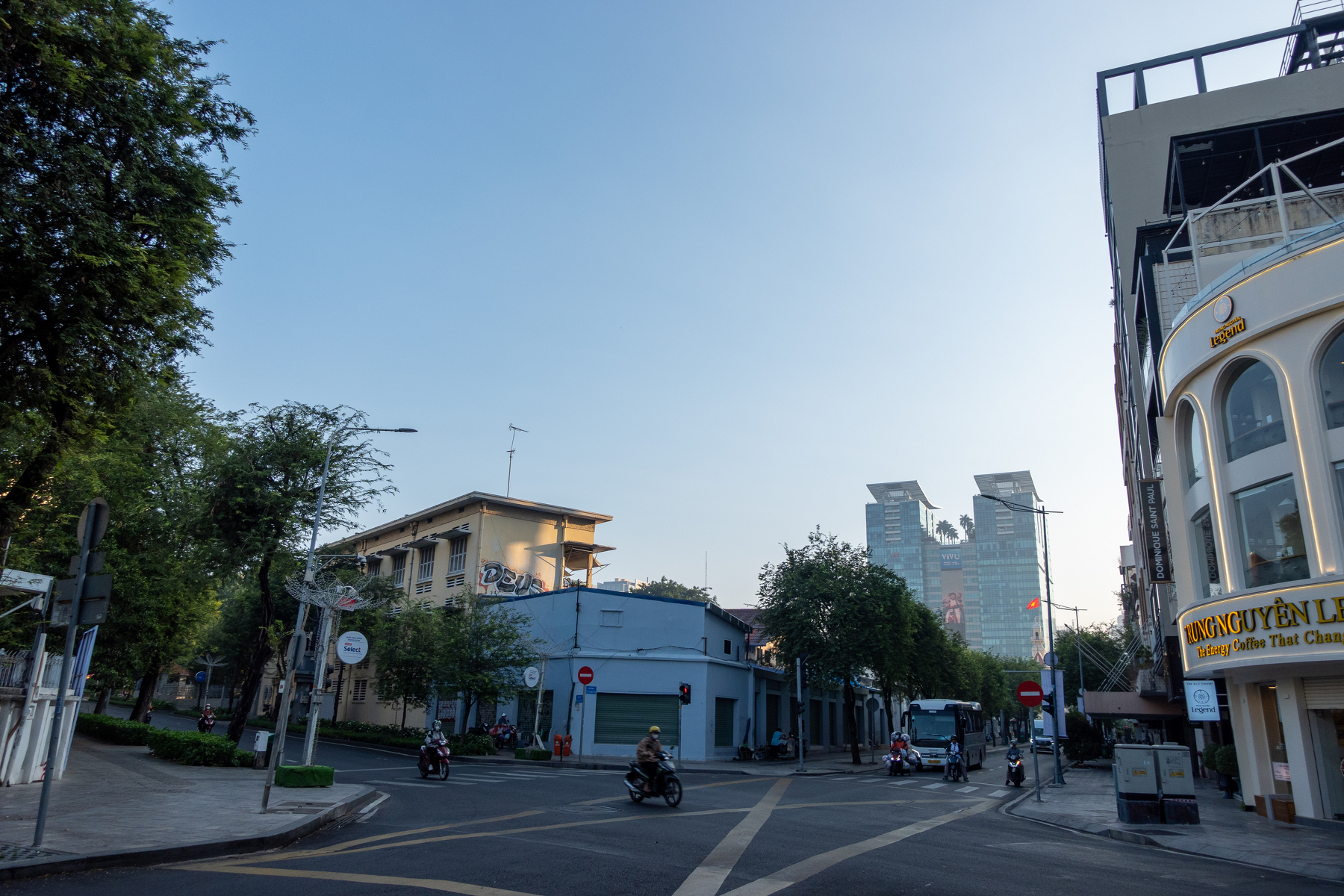 A quiet city street is lined with buildings and trees under a clear sky with a few motorcyclists riding along the road.