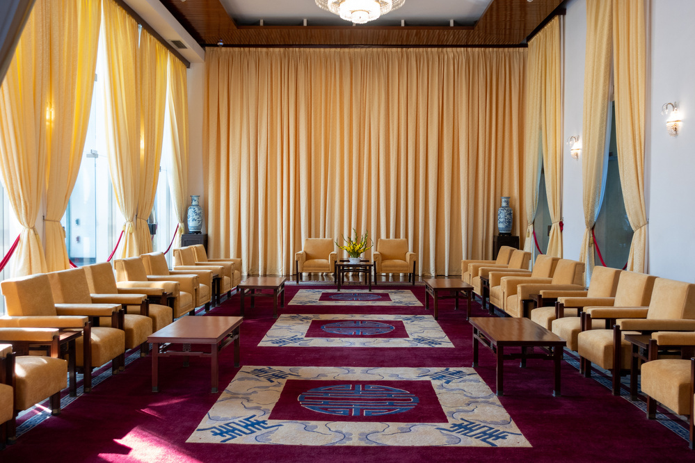 An elegant conference room features yellow curtains, beige armchairs arranged in rows, wooden tables, decorative vases, and a patterned carpet.