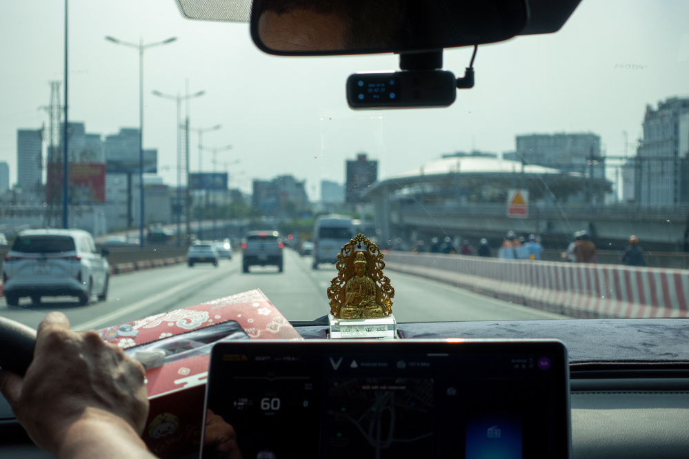A person is driving on a busy urban road with a small golden deity statue placed on the car dashboard.
